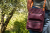 Person carrying a burgundy Karine leather bag outdoors with greenery in the background