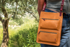 Person holding a brown leather crossbody bag outdoors with greenery in the background
